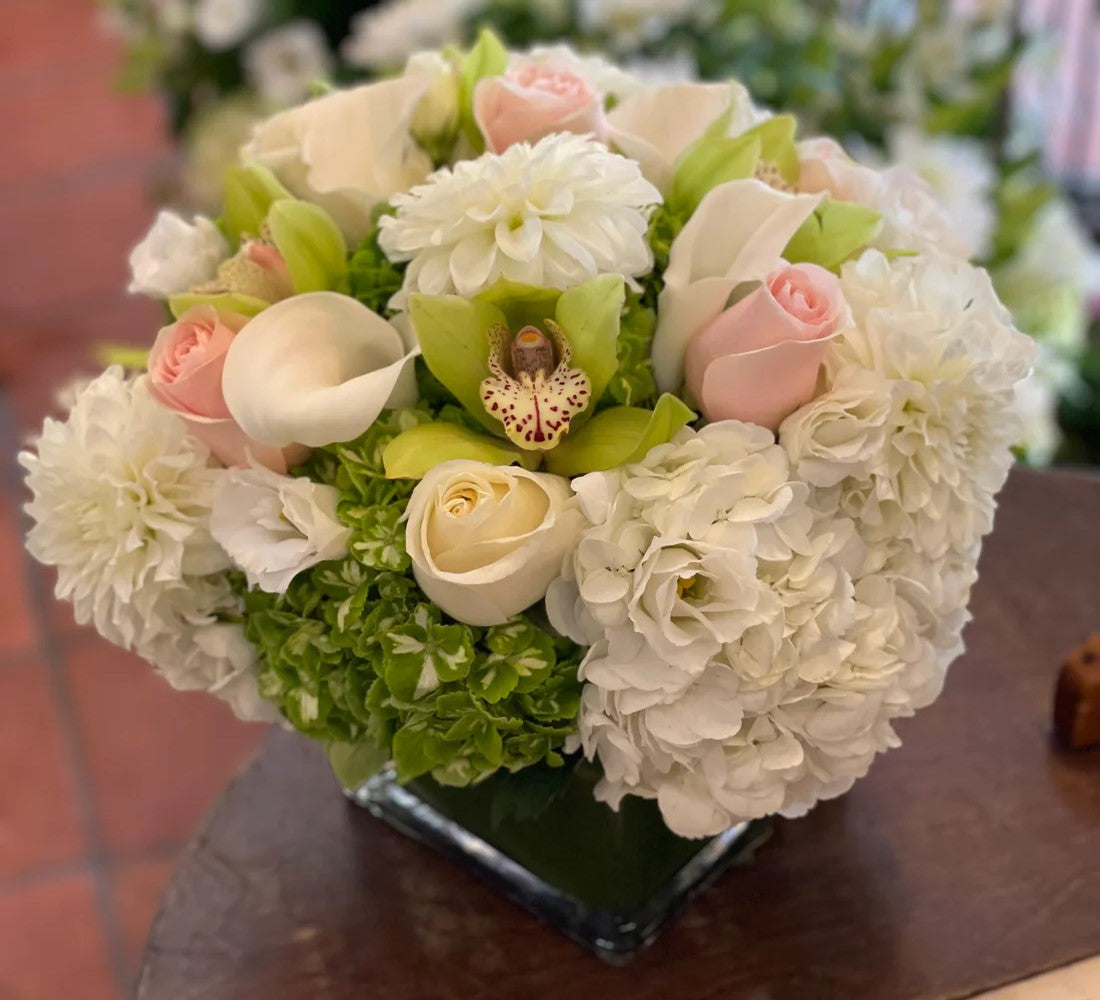 A floral arrangement featuring white and green Hydrangea, white calla lilies, pink roses, and cymbidium orchid heads in a square glass container. - Jerome Florists NYC