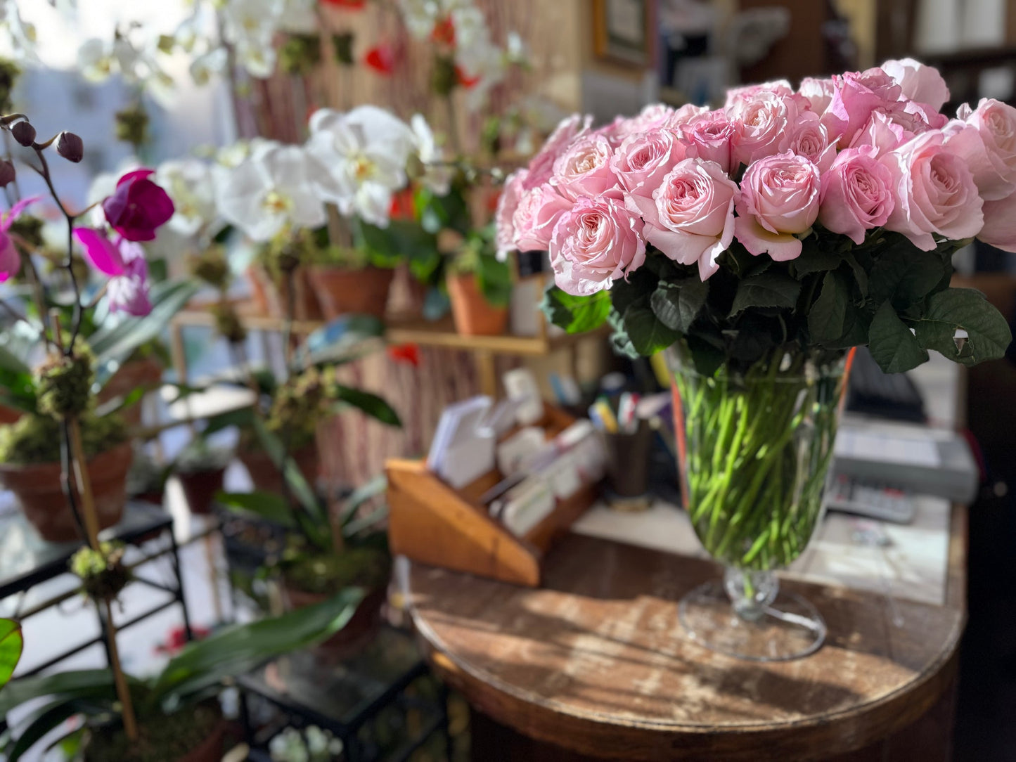 Pink roses in a glass vase beside white and purple orchids inside Jerome Florists' sunlit Upper East Side shop