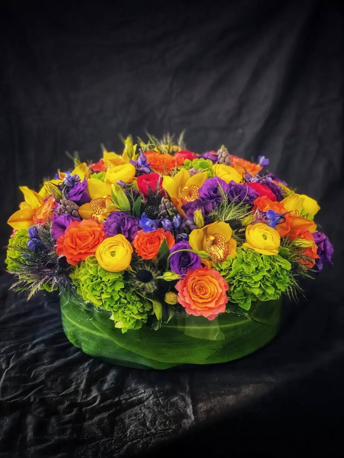 Colorful arrangement of orange roses, yellow ranunculus, orchids, and purple lisianthus in a leaf-wrapped base